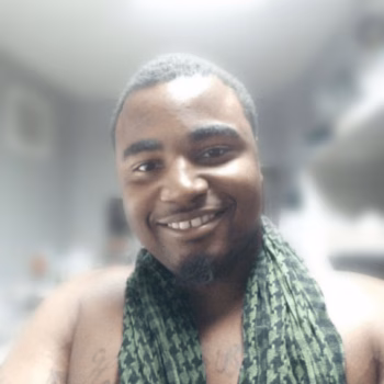 A smiling man with a green patterned scarf around his neck, shirtless, with tattoos on his chest.
