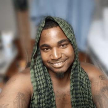 A smiling man with a green patterned cloth draped over his head and shoulders, showing tattoos on his arms.