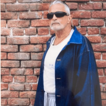 rm1 An older man with white hair and a beard, wearing sunglasses, a white t-shirt, and a blue denim jacket, stands against a red brick wall.