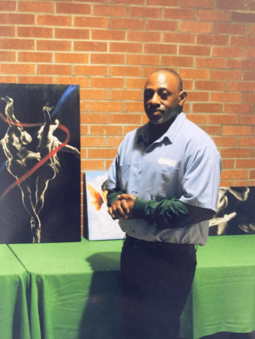 A man stands in front of a brick wall, next to a table displaying several paintings.