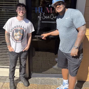 Two men standing in front of the HUMA of San Quentin entrance. The man on the left wears a Soundgarden t-shirt, and the man on the right, wearing a blue cap, holds the door handle.