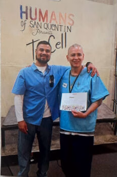 Edwin and a man in blue scrubs pose together in front of a 'Humans of San Quentin' banner, with Edwin holding a certificate.