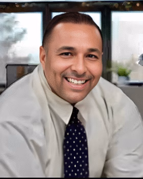 Jose Llaca, a man in a white shirt and patterned tie, smiles warmly at the camera in an office setting.