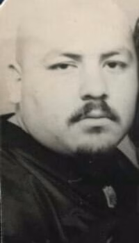 A black and white portrait of a bald man with a mustache and goatee, wearing a dark jacket with a small emblem.