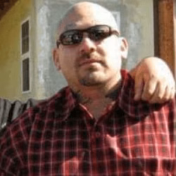 A bald man with sunglasses and a goatee wearing a red plaid shirt, with a hand on his shoulder.