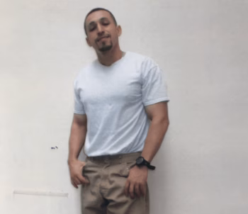 A man with a goatee wearing a white t-shirt and khaki pants, leaning against a plain wall.