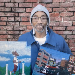 Kelvin, a man with glasses and a beanie, holds two paintings against a brick wall.