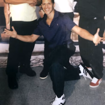 Paul 2 smiles and poses with arms outstretched, kneeling on a grey floor in front of other people.
