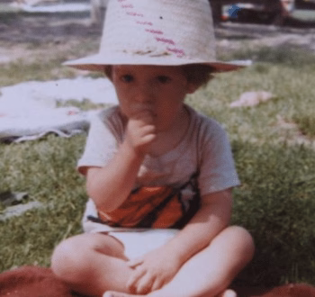 A young child wearing a wide-brimmed straw hat sits cross-legged on a red blanket in a grassy field, with a finger near their mouth.