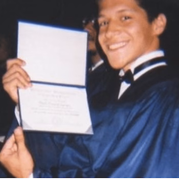 Paul 4 smiles while holding a diploma in a blue graduation gown.