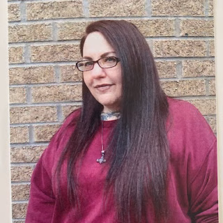 Sarah 2 Sarah-2, a woman with long dark hair and glasses, wearing a purple shirt and a necklace, stands in front of a brick wall.