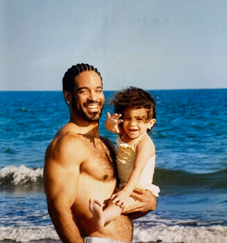A smiling man named Terence holds a young child on a sunny beach with the ocean in the background.