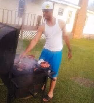 A man in a white tank top and blue shorts grilling meat on a black barbecue in a backyard.