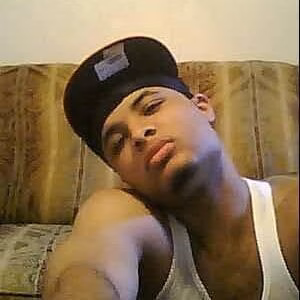 A man wearing a black baseball cap and a white tank top resting his head on his arm while sitting on a couch.