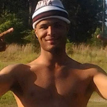 A shirtless man wearing a striped beanie with 'Trell' written on it, smiling and making peace signs with both hands in an outdoor setting.