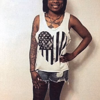 ebony 3 A woman with glasses and tattoos on her arms stands against a wall, wearing a white tank top with a heart and American flag design.