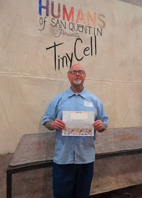 eric b 1 Eric B, 62, stands in front of a banner for 'Humans of San Quentin Presents TinyCell' holding a certificate.