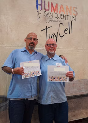 eric b 3 Two men in light blue shirts hold certificates in front of a wall with the text 'HUMANS of SAN QUENTIN Presents TinyCell'.