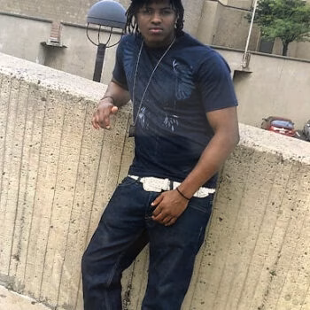 A man with dreadlocks wearing a blue 'SoulDawn' cap, a dark t-shirt, and jeans, leaning against a concrete wall.