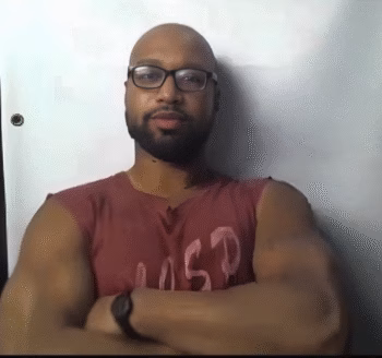 A bald man with glasses and a beard, wearing a red sleeveless shirt, stands with his arms crossed against a white wall.