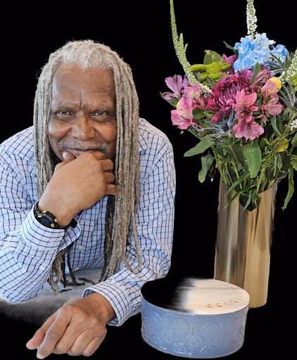 Gerald, a man with long grey dreadlocks, rests his chin on his hand while looking at a floral arrangement in a gold vase and a decorative box.