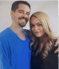 A man with a beard in a blue shirt and a blonde woman in a black top posing together.