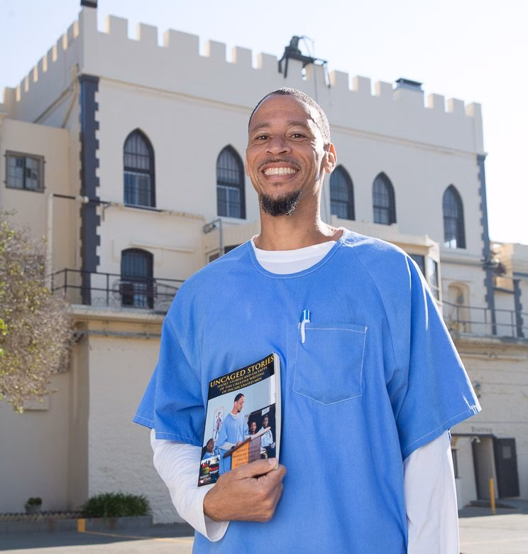 A smiling man in blue scrubs holds a book titled 'Uncaged Stories' in front of a historic white building.