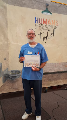 A man with a beard and glasses in blue scrubs holds a certificate in front of a mural for Humans of San Quentin Presents TinyCell.