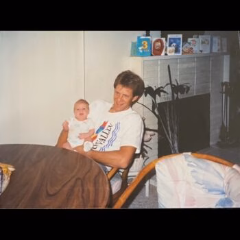 Man holding baby A man in a white t-shirt holds a baby in a room with a fireplace and cards on the mantel.