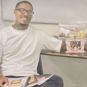 A smiling man with glasses and neck tattoos sits at a desk, holding a magazine and resting his arm near a heart-shaped photo frame and a bag of groceries.