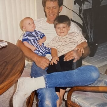 Man with two children A man in a white t-shirt and blue jeans sits in a chair, holding a baby in a blue and white striped onesie and a young boy in a striped shirt.