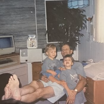 Family moment in bedroom A man lies on a bed with two young children, one on his chest and another on his lap, in a bedroom with a computer and a window.