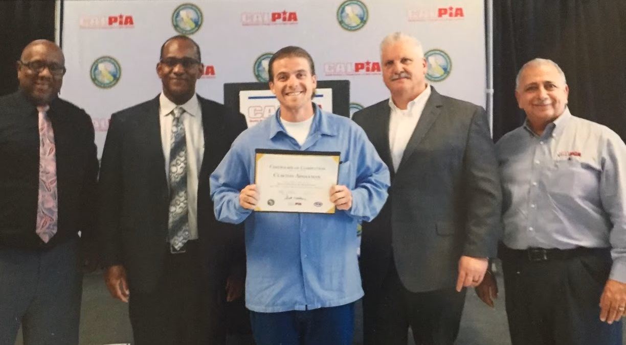 Five men stand in front of a CALPIA backdrop. The man in the center, wearing a blue work shirt, holds a certificate of completion.