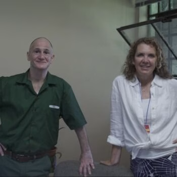 A bald man in a green shirt and a woman with curly hair in a white shirt stand side-by-side in an indoor setting.