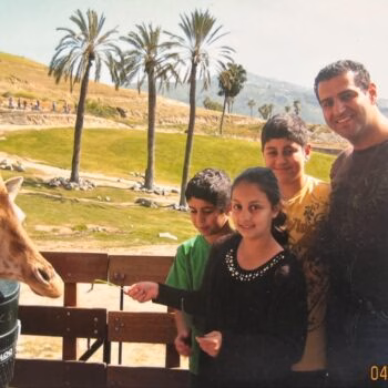 A family of four, including a man and three children, stands on a wooden platform feeding a giraffe. Palm trees and a grassy hill are visible in the background under a clear sky.