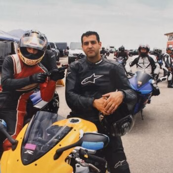 A man in a black racing suit sits on a yellow motorcycle in front of a group of other motorcyclists at a track, with a building labeled 'JOHN HARTMAN GARAGE' in the background.