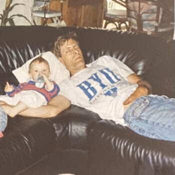 Man and baby on couch A man in a white BYU shirt and a baby in a red and blue outfit are resting on a black leather couch.