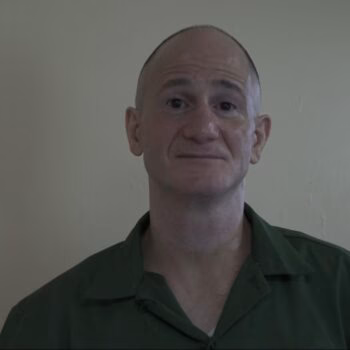 A bald man with a slight smile, wearing a green shirt, against a plain wall.