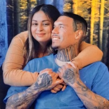 A woman with long dark hair embraces a man from behind. The man, wearing a blue shirt, has extensive tattoos on his neck and arms, including a '100' on his hand. They are smiling and looking at each other against a forest backdrop.