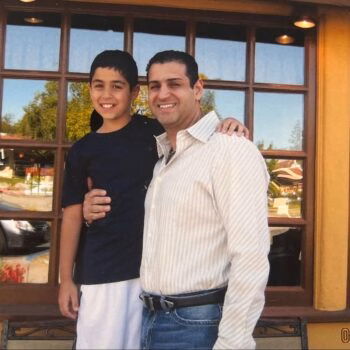 A man and a young boy stand together smiling in front of a building with large windows.