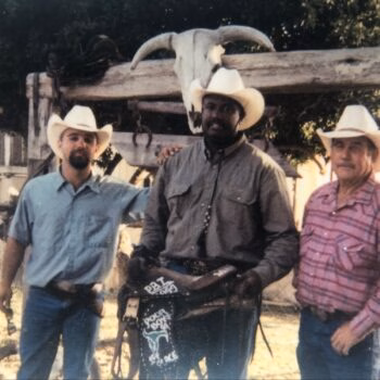 Three men in cowboy hats stand in front of a wooden structure with a bull skull mounted on top.