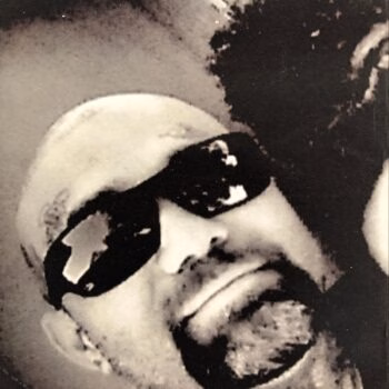 Close-up of a person's face wearing dark sunglasses, captured in a grainy black and white photograph.