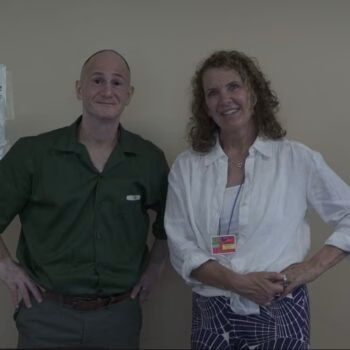 A man and a woman stand side-by-side in front of a wall with several papers taped to it.