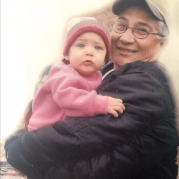 A smiling older man in a cap and glasses holds a baby in a pink outfit and hat.