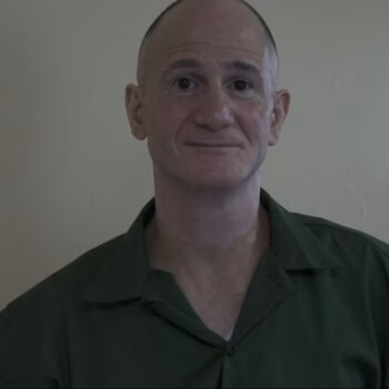 A bald man with a slight smile wearing a green shirt against a plain wall.