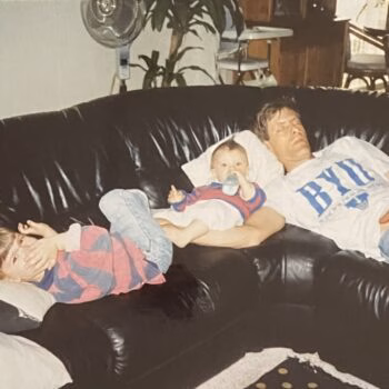 Man and children on couch A man wearing a BYU shirt lies on a black leather couch with two young children, one sleeping on his chest and another resting his head on his shoulder.