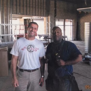Two men standing in a construction site. The man on the left wears a white t-shirt with a logo, and the man on the right wears a dark blue shirt with a tool belt.
