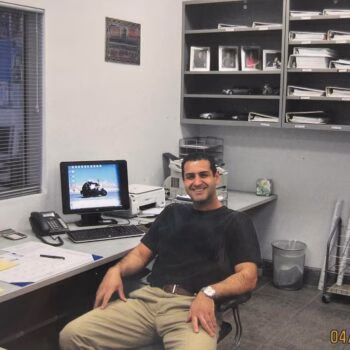 A man sits in an office chair at a desk with a computer, surrounded by shelves of documents and a window.