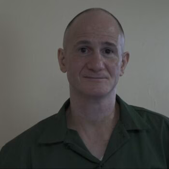 A bald man with a slight smile wearing a green shirt, looking directly at the camera against a plain wall.
