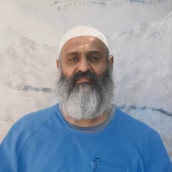 A man with a full grey beard and a white skullcap, wearing a blue shirt, stands in front of a mountain landscape.